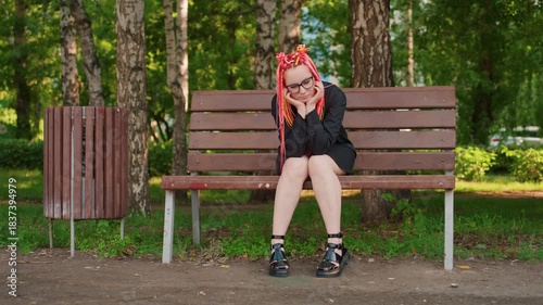 full body frontal shot of woman with bright dreadlocks seated on bench between trees, hands clasped under chin, boots and casual dress visible, summer park atmosphere, reflective posture