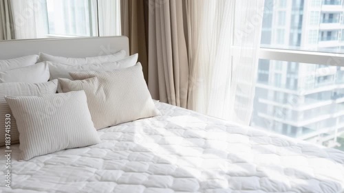 A white bed with a white comforter and white pillows. The bed is in a room with a large window that lets in a lot of light