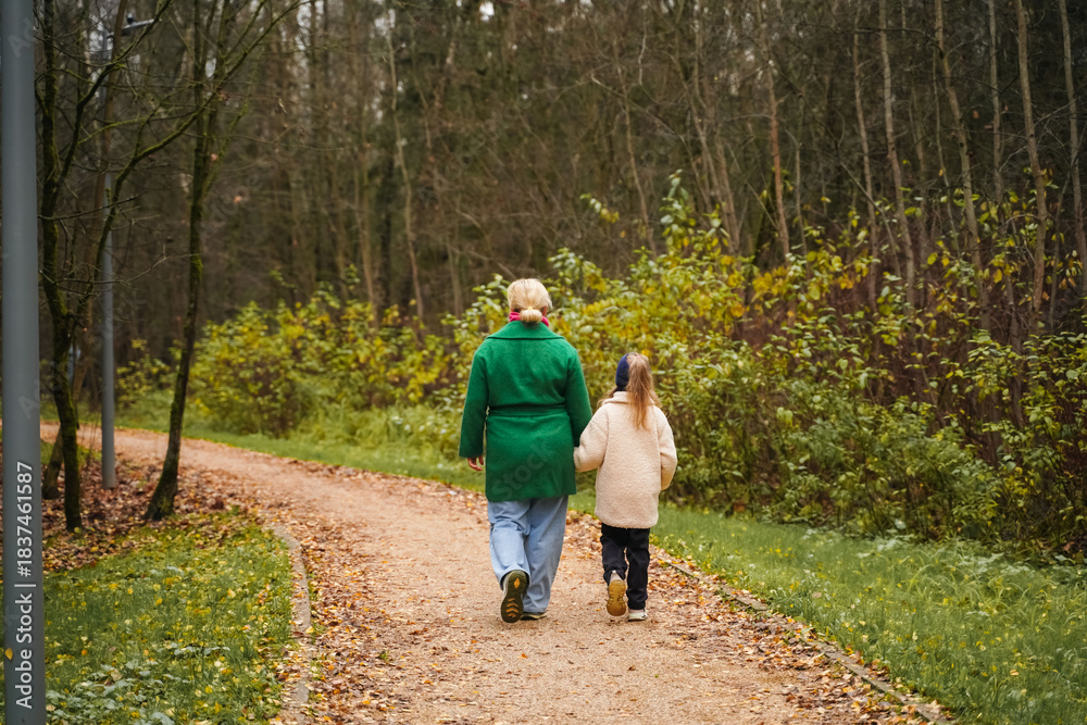 Fototapeta premium Walking mother with daughter on park path. Holding hands shows parenting, care, trust, safety. Outdoor family activity supports bonding, happiness, healthy lifestyle, weekend leisure, forest trail.