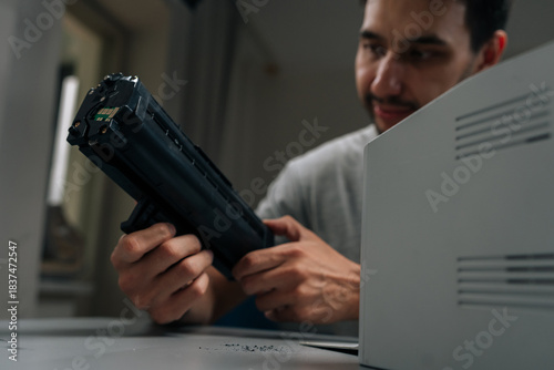 Close-up portrait of bearded repairman smiling performing printer maintenance, replacing empty toner cartridge with new one on desk, ensuring smooth office operations and diy repair.