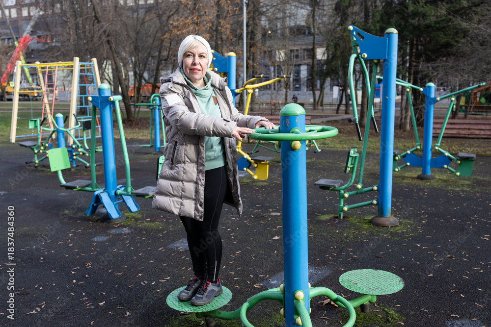 Fototapeta premium A woman with short hair is working out on outdoor exercise equipment in a park. She is wearing a light jacket and black trousers.
