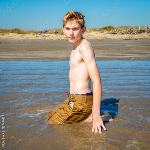 Shirtless young boy at the beach sitting in the water, having fun playing in the mud and sand at his feet