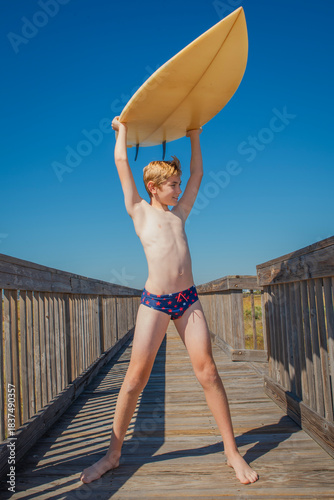 Smiling boy in swim suit carrying his surfboard over his head on a walkway near the beach