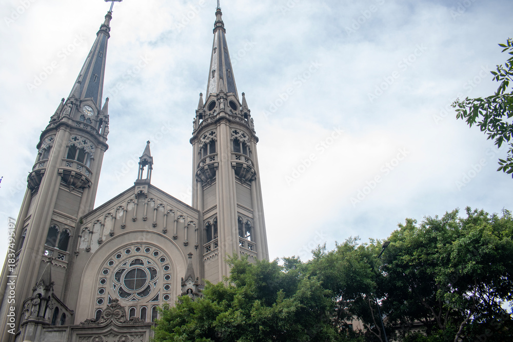 Obraz premium Majestic gothic church façade with tall twin towers, intricate architectural details, and a large rose window rising above lush green trees under a bright cloudy sky