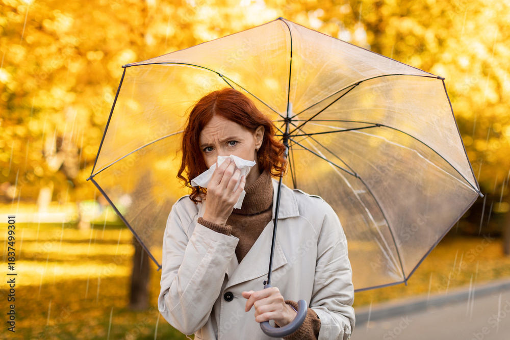 Naklejka premium A woman with red hair is standing in a park, holding a tissue while she uses a clear umbrella for protection from the rain. Autumn leaves are falling around her, adding color to the scene.