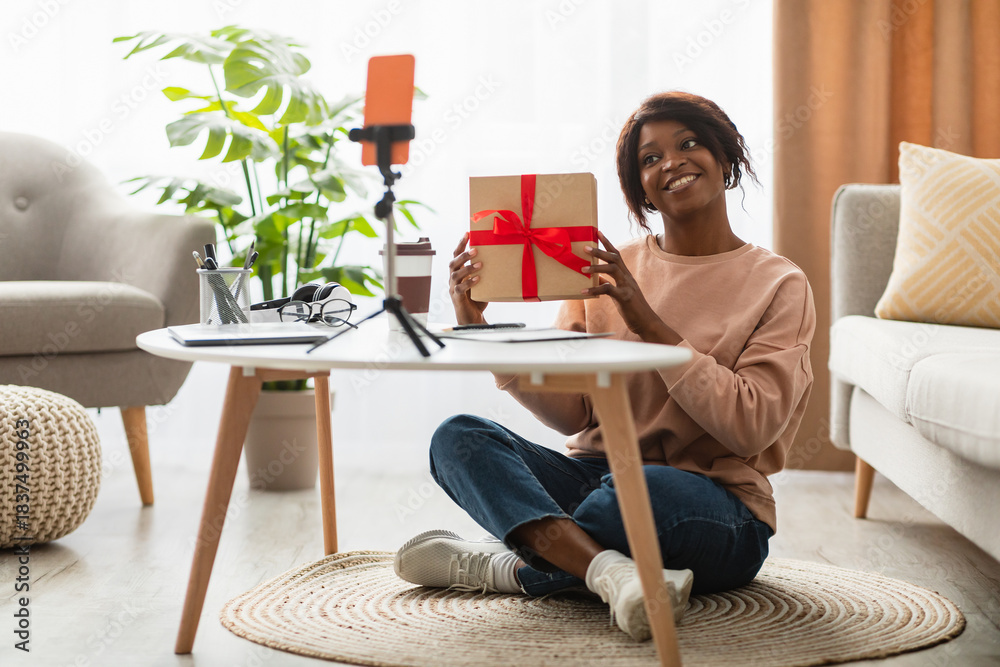 Naklejka premium A cheerful woman sits on the floor in her living room, holding a gift wrapped with a red ribbon. She is recording an unboxing video with a smartphone on a tripod.