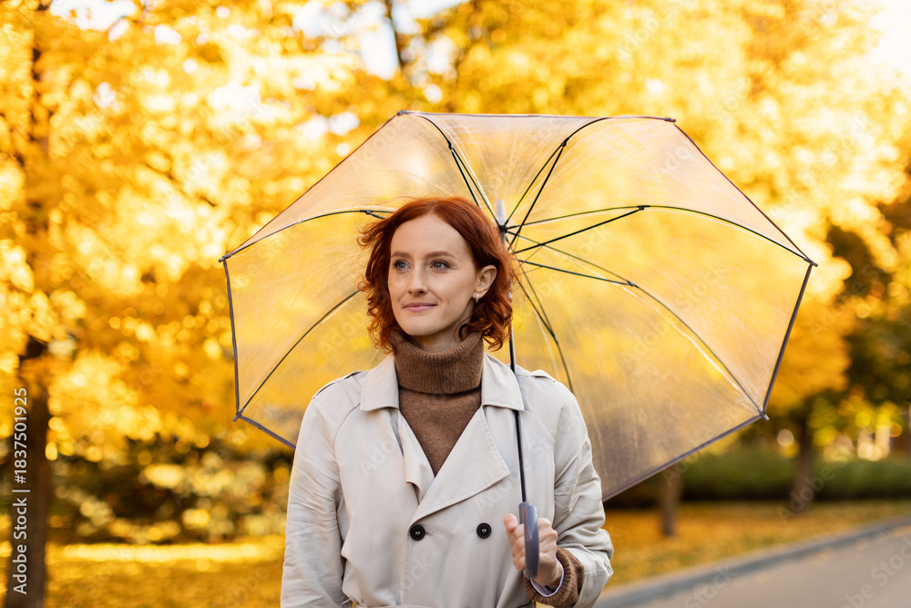 Naklejka premium A woman with red hair enjoys a walk in a park filled with yellow autumn leaves. She holds a clear umbrella while wearing a beige coat, smiling at the pleasant weather.