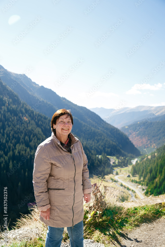 Naklejka premium Active smiling senior woman, happy elderly traveler standing against scenic Fagaras mountains landscape. Transfagarasan Highway, Romania. Travel concept