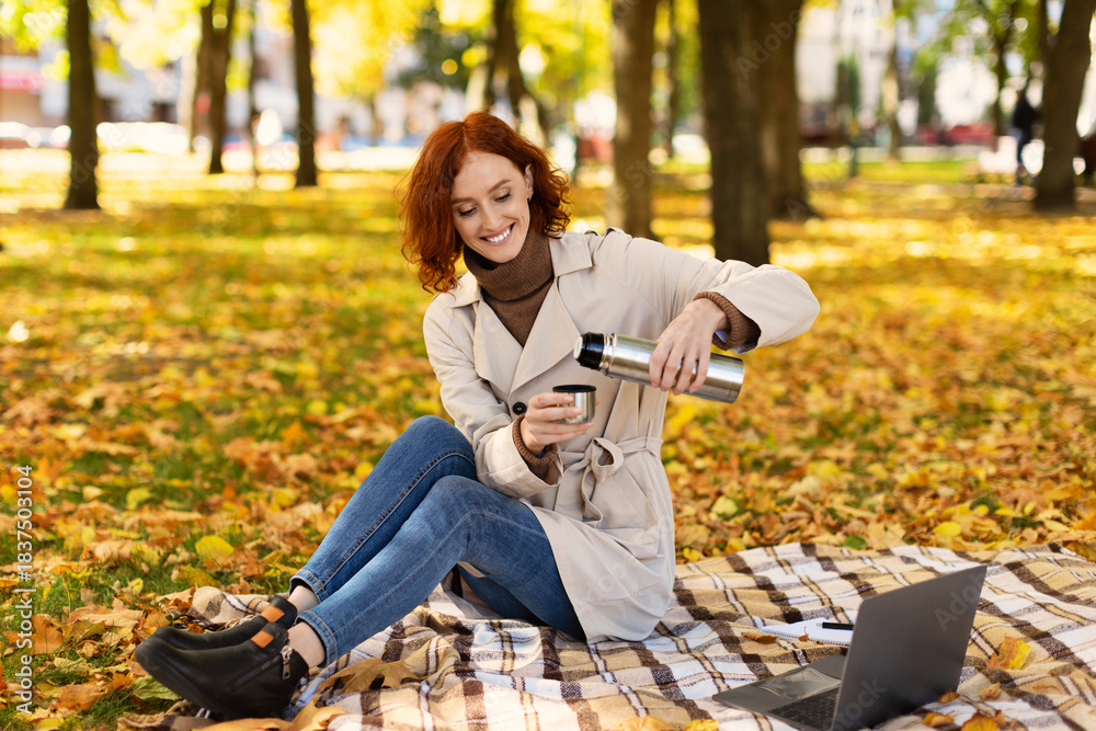 Naklejka premium A woman sits on a blanket in a park, pouring a warm drink from a thermos into a cup. The ground is covered in colorful autumn leaves, creating a picturesque scene.