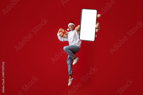A young man wearing a Santa hat happily jumps while holding a smartphone in one hand and a gift in the other. The vibrant red background adds to the festive atmosphere.