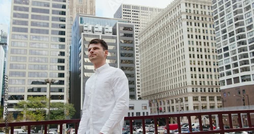 A young successful brunette businessman dressed in a white shirt folds his arms in front of him against the backdrop of downtown and looks into the camera.