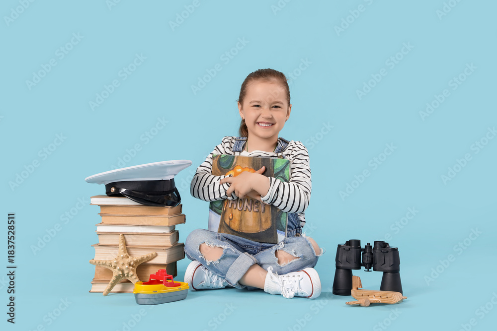 Fototapeta premium Cute little girl with adventure books and toys sitting on blue background