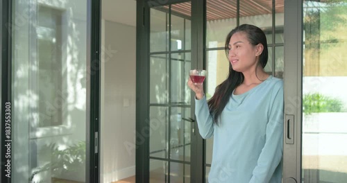 Mid adult Asian woman standing on sliding glass threshold savoring herbal tea while gazing yardward