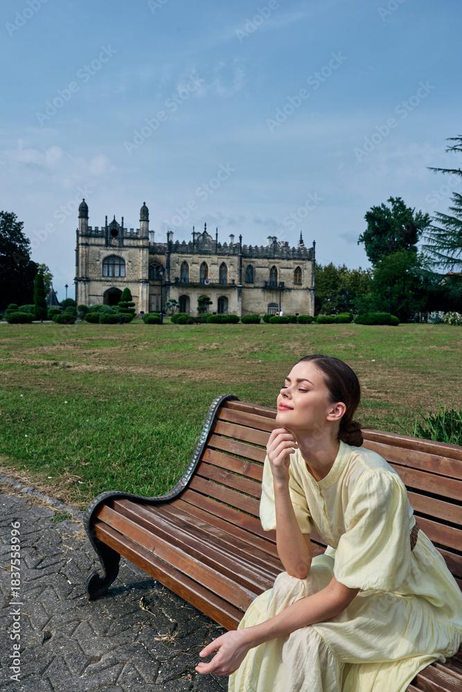 Naklejka premium Woman on bench in park with historic mansion background, relaxation in sunlight, authenticity inclusivity