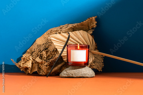 Red candle with flame placed on stone with dried palm leaf and textured paper on blue and orange background. Aromatherapy and cozy home atmosphere concept. Mockup