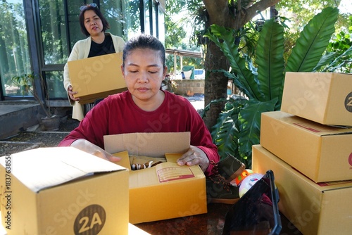 A woman is intently packing an item into a package box, with another woman standing behind her holding a box.
