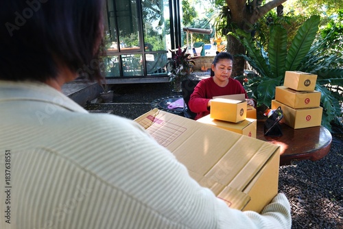 A view from behind of a woman holding a box, with another woman sitting in front of her surrounded by package boxes.