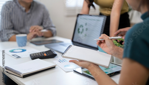 Close-up of a professional holding a blank notebook and stylus, taking notes during a team meeting. Unrecognizable colleagues discuss data in the background.