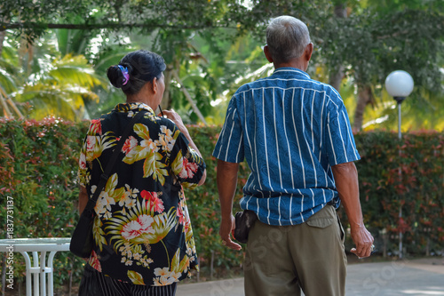 A rear view of an elderly couple walking down a paved path in a garden, the man wears a striped shirt and khaki pants, while the woman in a floral shirt walks beside him toward greenery.