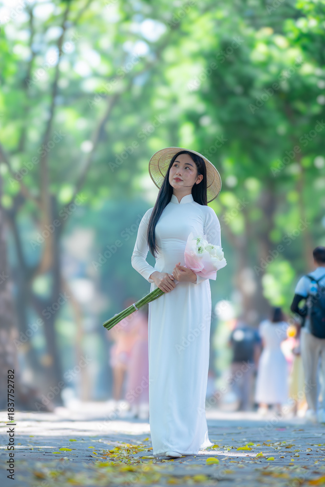 Obraz premium A young Vietnamese woman wearing a traditional white ao dai and a conical hat smiles gracefully under bright sunlight, on street in Hanoi, Vietnam.