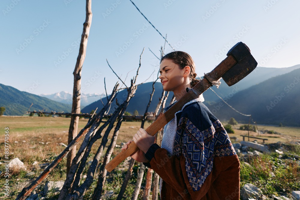 Obraz premium Woman, axe, countryside, fence, mountains, field, smiling, outdoors carrying an axe on her shoulder near a rustic wooden fence in a sunny mountain valley, rural portrait with confident expression.