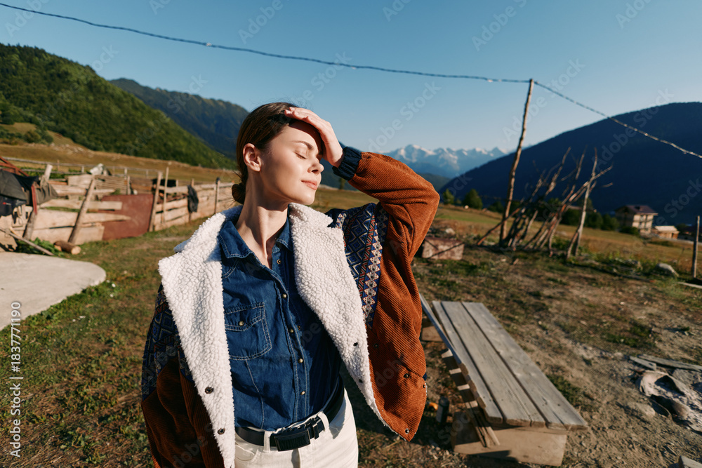 Obraz premium Woman portrait in countryside mountains with sunlight on face, relaxed outdoor fashion look wearing cozy jacket and denim shirt near wooden bench and rustic fence in scenic nature.