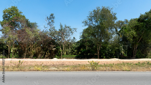 Horizontal view of dry asphalt road. Parallel road behind is a road under construction. Blocked by grass and ditches. Background of trees forest under blue sky.