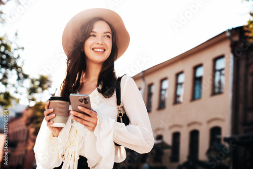 Modern young woman walking in the street. Model texting and holding cup of coffee. Businesswoman holds smartphone and looking away outdoors. Beautiful female in the city in sunny day in hat, outdoors