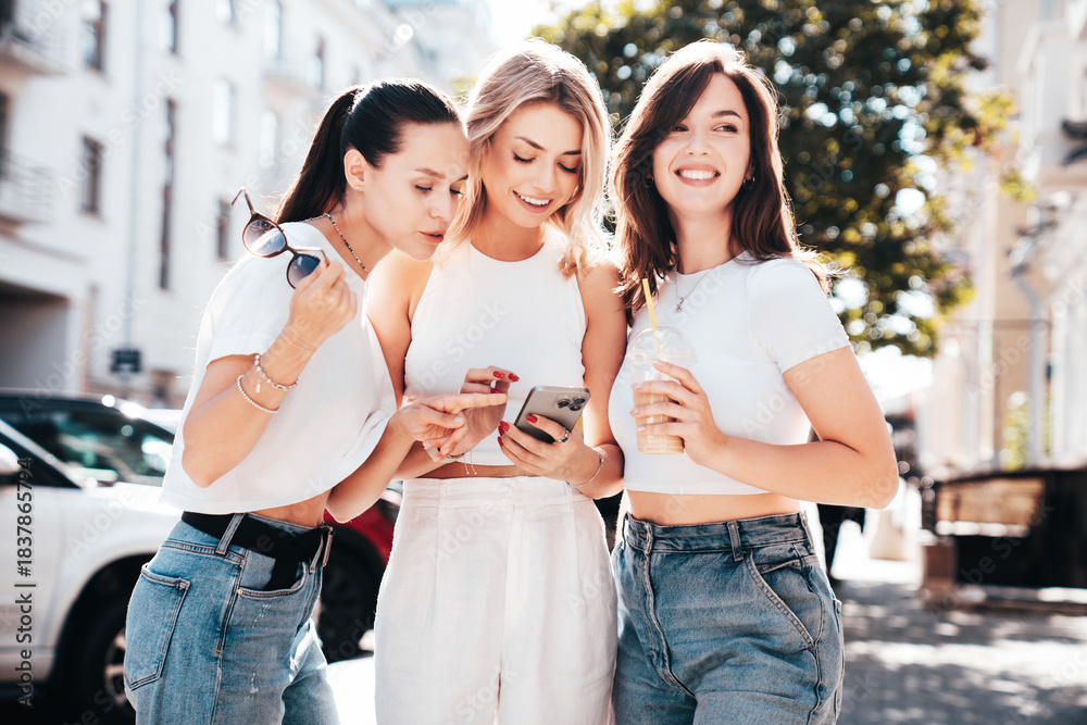 Fototapeta premium Three young beautiful smiling trendy female. Carefree women posing in street. Positive models having fun. Cheerful and happy. Hold smartphone, look at mobile cellphone screen, use phone apps