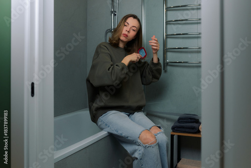 Woman brushing her hair while sitting on the edge of a bathtub in a modern bathroom
