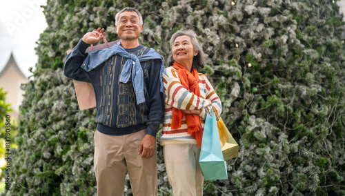 Happy senior asian couple holding shopping bag in front of snowy Christmas tree, enjoying winter holiday season, new year end sale festive shopping, senior lifestyle, love, joy and holiday celebration