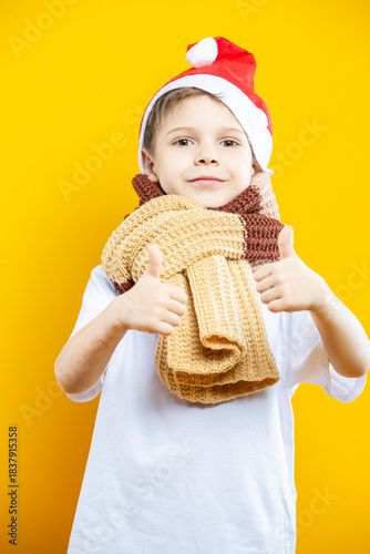Portrait of Preschooler Caucasian Boy In Santa Hat and Warm Knitted Scarf With Lifted Hands Thumbs Up Isolated On Yellow Background as Image To Convey New Year and Christmas Preparation Ideas