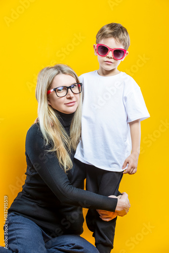 Young Smiling Cheerful Happy Parent Couple Woman And Child Boy In Casual Clothing and Glasses Having Mommy Little Kid Son Hug Cuddle Looking Straight on Yellow Background