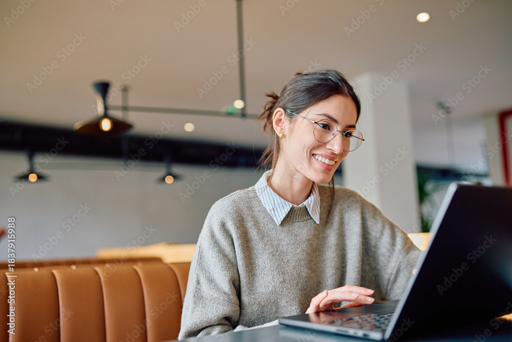 © we.bond.creations - Young smiling woman wearing glasses using laptop at a modern cafe, enjoying remote work and online learning experience