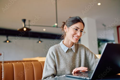 Принти на полотні Young smiling woman wearing glasses using laptop at a modern cafe, enjoying remo