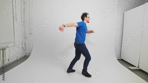 Side view of cheerful Caucasian guy in T-shirt and trousers jumping and moving filmed in slow motion. Young adult bearded guy hops showcasing fun and energy in studio with white walls. Movement theme.