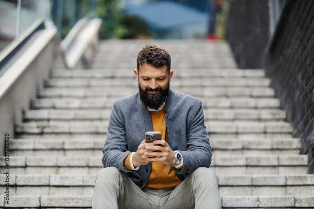 Obraz premium Portrait of smiling businessman sitting on stairs with cellphone in hands and texting messages at financial district downtown.