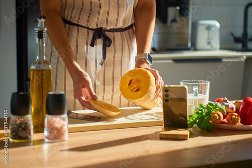 Person in apron holding sliced butternut squash on cutting board in kitchen. Woman slicing pumpkin on table with olive oil, spices, vegetables and smartphone. Concept of cooking and healthy food