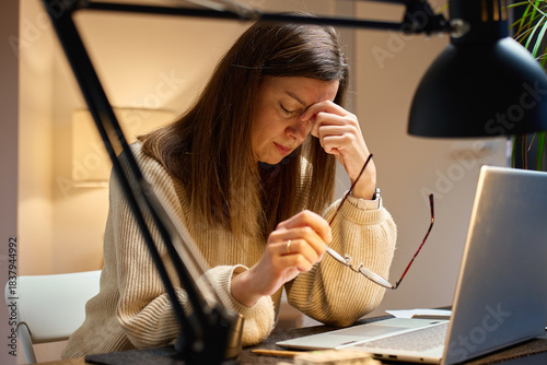 Tired woman sitting at desk with laptop, rubbing eyes while holding glasses in hand. Fatigued person working late. Concept of eye strain, stress, headache and overwork