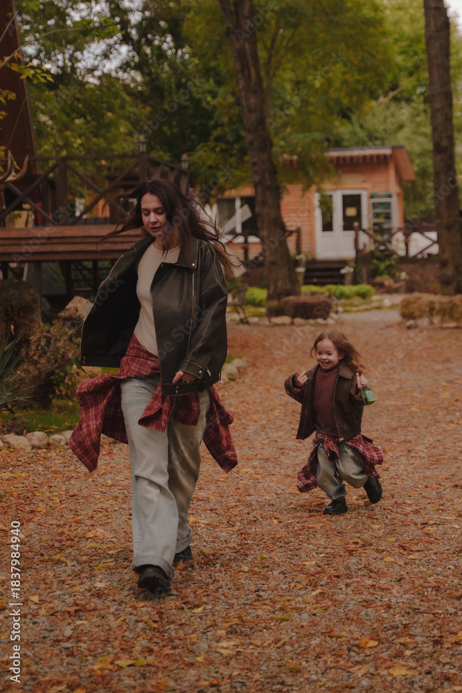 Obraz premium Mother and daughter run around the autumn yard near cozy A-shaped houses. The girls appear to be dressed in leather jackets, blue jeans and plaid shirts. Autumn atmosphere. Vertical photo.