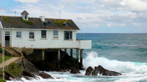 White building on stilts overlooking a turbulent ocean with waves crashing against rocks.