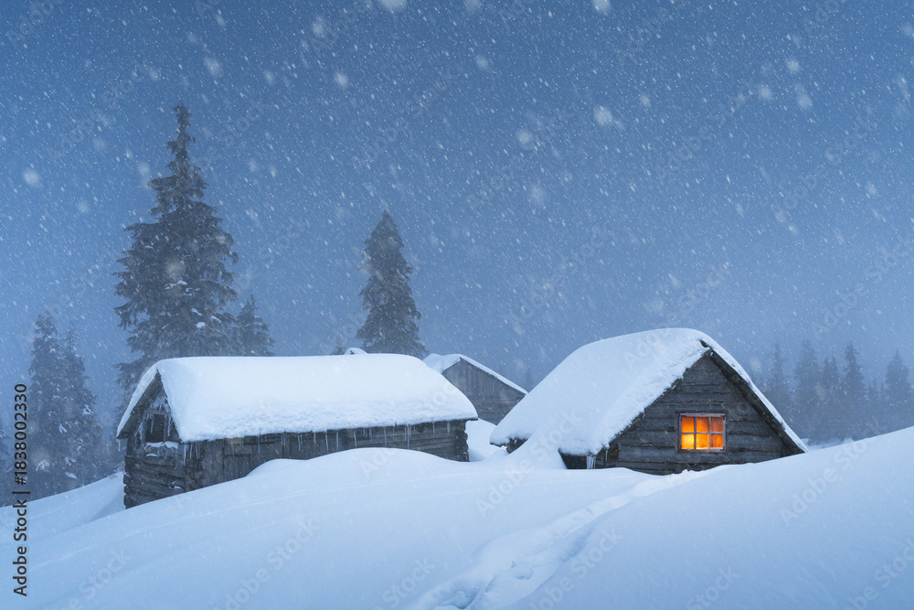 Fototapeta premium Cozy house with warm light during winter snowfall in a snowy forest. Cold mountain night landscape with snow covered trees