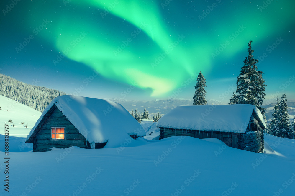 Fototapeta premium Aurora Borealis over snowy mountain landscape with a small cozy house. Colorful Northern lights in winter night nature
