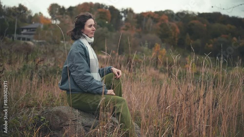On a rock, a beautiful white European woman with Slavic features, dressed in a light blue jacket and white scarf, sits peacefully in a serene autumn landscape filled with warm colors.