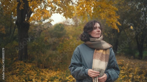 Yellow leaves, crowns of trees A beautiful Slavic girl in a cozy scarf stands in a serene outdoor setting, surrounded by vibrant autumn trees, reflecting on nature with a gentle smile.