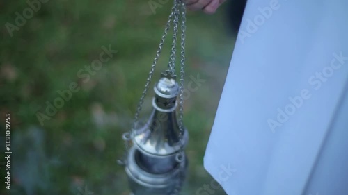 Close-up of a silver metal thurible or censer hanging from chains, actively swinging and emitting thick incense smoke during a religious or commemorative service outdoors.
