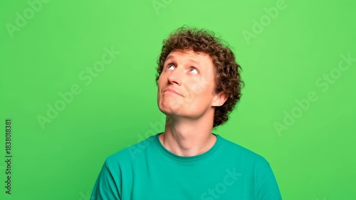 Young man male adult curly hair wearing teal green shirt looking upward thoughtfully against bright green background