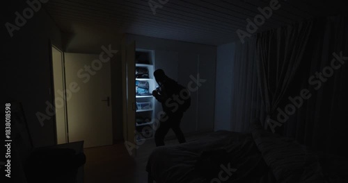 A masked intruder moves through a dark bedroom using a flashlight to search shelves and drawers, symbolizing burglary, home invasion, theft, and nighttime danger.