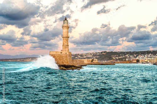 Old Venetian Lighthouse in Chania Crete hit by powerful turquoise wave under dramatic pastel sky. Scenic harbor view with dynamic sea motion, stone pier, warm evening light, stormy coastal atmosphere.