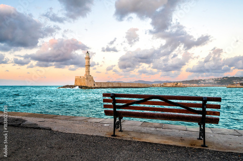 Wooden bench overlooking Venetian Lighthouse in Chania Crete Greece during calm evening with pastel clouds, turquoise sea, peaceful harbor scenery, tranquil coastal mood, inviting Mediterranean atmosp